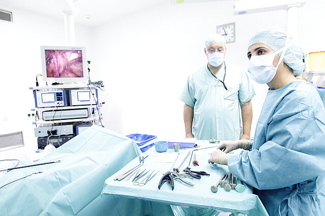 Ein Mann und eine Frau in Operationsbekleidung mit Mundschutz und chirurgischen Instrumenten stehen in einem Operationsraum
