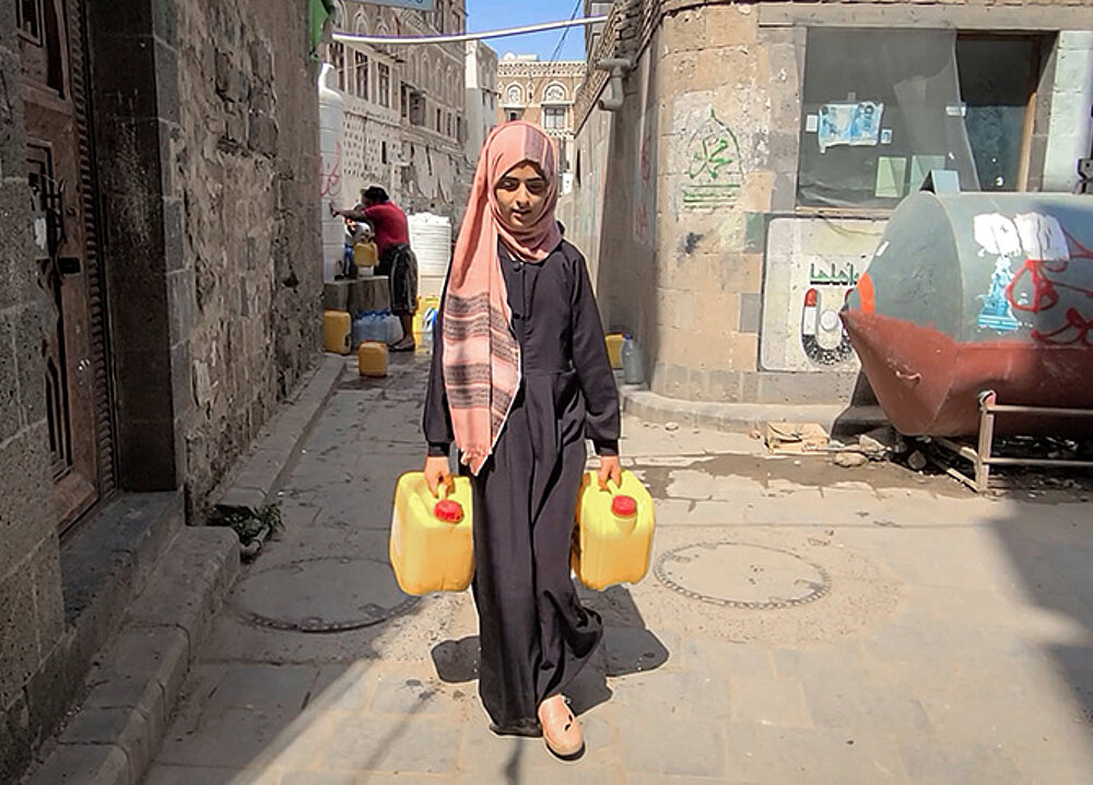 Ein Mädchen läuft eine Straße entlang und trägt zwei gelber Wasserkanister