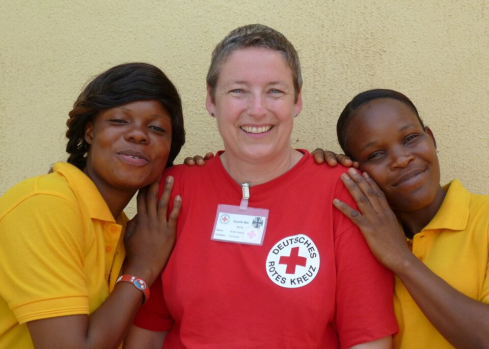 Eine DRK-Mitarbeiterin im roten Shirt mit DRK-Logo wird umrahmt von zwei liberianischen Kolleginnen