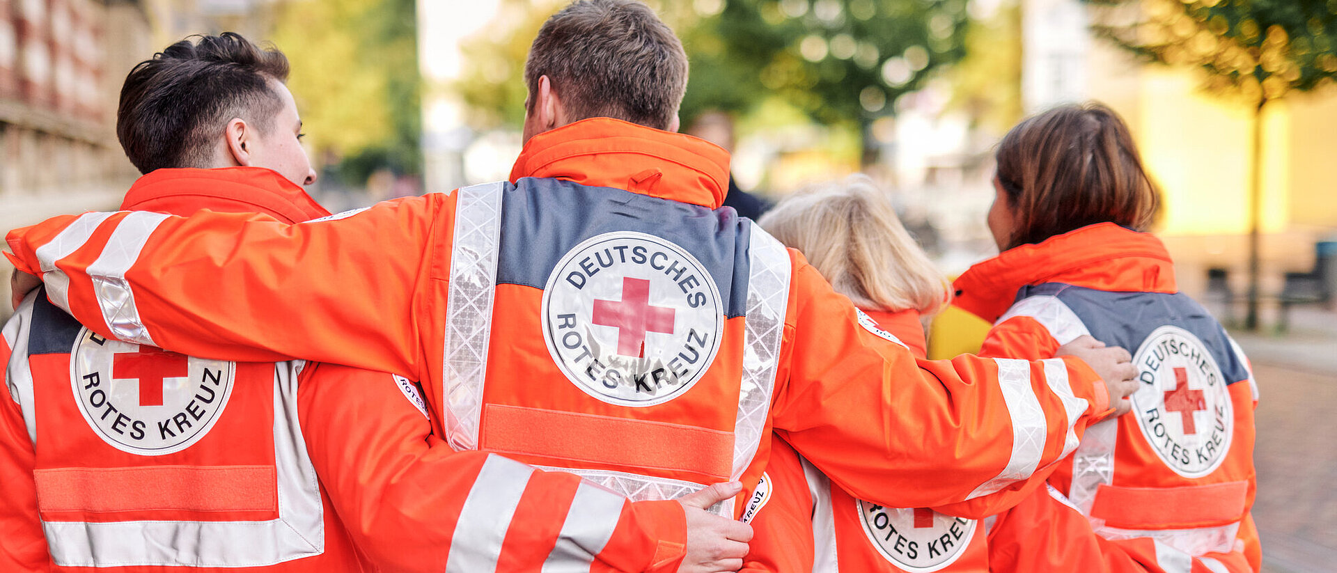 Vier Personen, die leuchtend orangefarbene Rettungswesten des Deutschen Roten Kreuzes tragen, stehen mit verschränkten Armen in einer städtischen Umgebung. Auf den Rücken der Westen sind das Logo und der Schriftzug "Deutsches Rotes Kreuz" deutlich sichtbar. Die Gruppe zeigt Solidarität und Zusammenhalt.