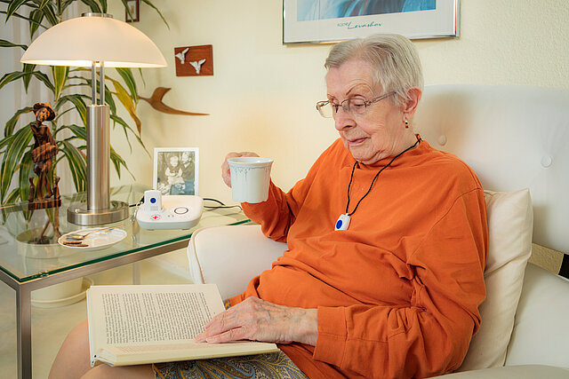 Eine Seniorin sitzt und liest ein Buch. Sie trägt eine Kette mit dem Hausnotruf-Knopf um den hals