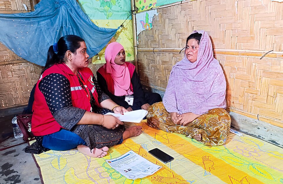 Drei Frauen sitzen auf dem Boden einer Hütte. Zwei davon tragen rote Westen.