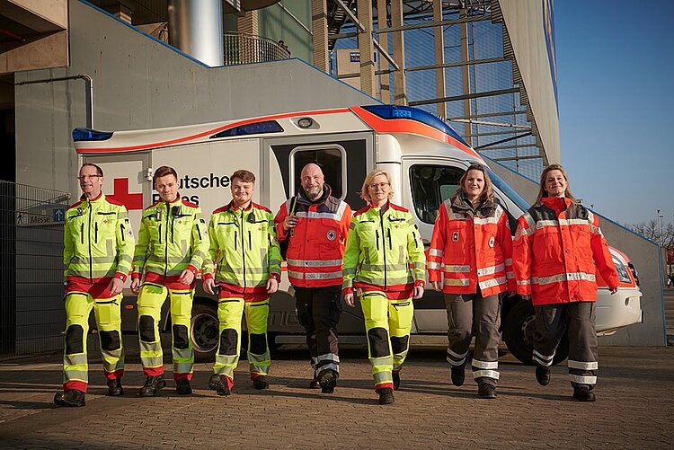 Hauptamtliche und ehrenamtliche Sanitäter und Sanitäterinnen und Einsatzleiter vor Ihrem Rettungswagen am Stadion