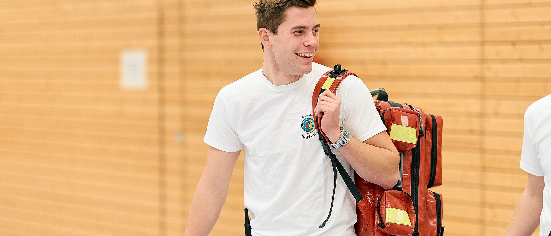 Helfer vom Deutschen Jugendrotkreuz Ein junger Mann mit JRK-T-Shirt hält einen Erste-Hilfe-Rucksack über der Schulter und lächelt