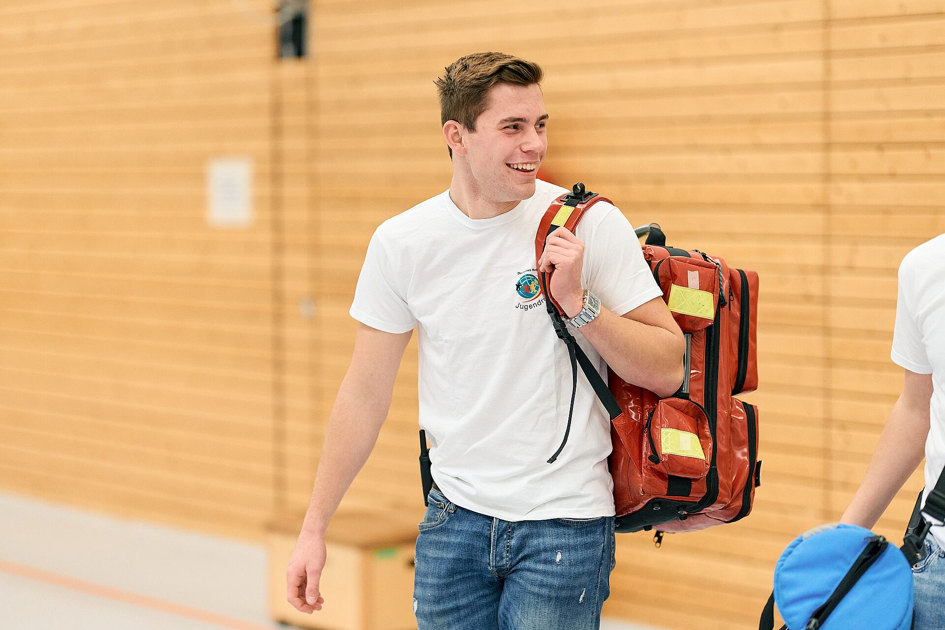 Ein junger Mann mit JRK-T-Shirt hält einen Erste-Hilfe-Rucksack über der Schulter und lächelt