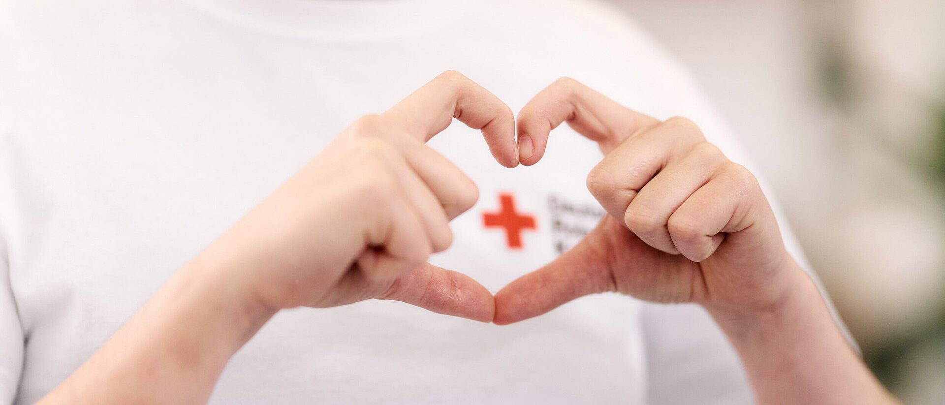 Das Bild zeigt den Oberkörper einer Person, ihr Gesicht ist nicht zu sehen. Diese Person formt mit den Daumen und Zeigefingern beider Hände ein Herz, welches das Rote Kreuz umrahmt, welches auf das weiße T-Shirt der Person gedruckt ist.