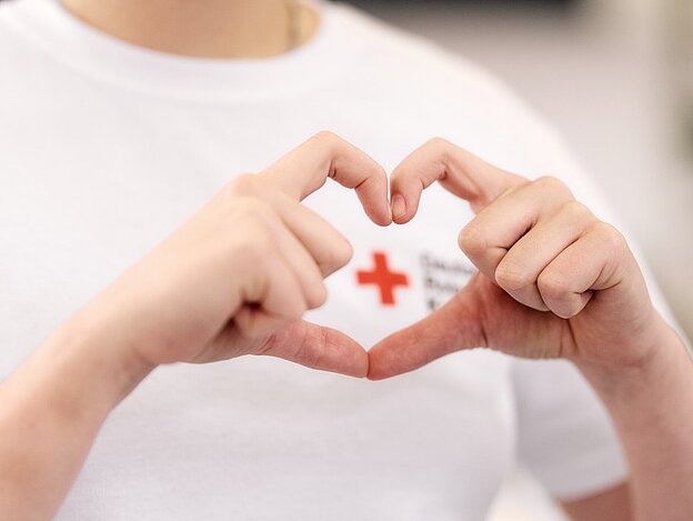 Das Bild zeigt den Oberkörper einer Person, ihr Gesicht ist nicht zu sehen. Diese Person formt mit den Daumen und Zeigefingern beider Hände ein Herz, welches das Rote Kreuz umrahmt, welches auf das weiße T-Shirt der Person gedruckt ist.