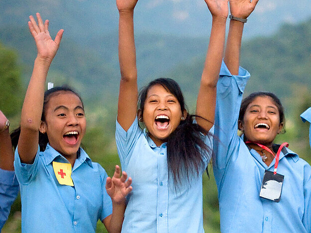 Zu sehen sind sechs Mädchen, die jubelnd und lachend in einer Reihe stehen. Sie alle tragen blaue Hemden, die Teil einer Schuluniform sind. Im Hintergrund erkennt man bewaldete Hügel.