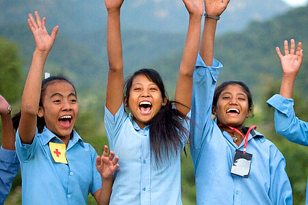 Zu sehen sind sechs Mädchen, die jubelnd und lachend in einer Reihe stehen. Sie alle tragen blaue Hemden, die Teil einer Schuluniform sind. Im Hintergrund erkennt man bewaldete Hügel.
