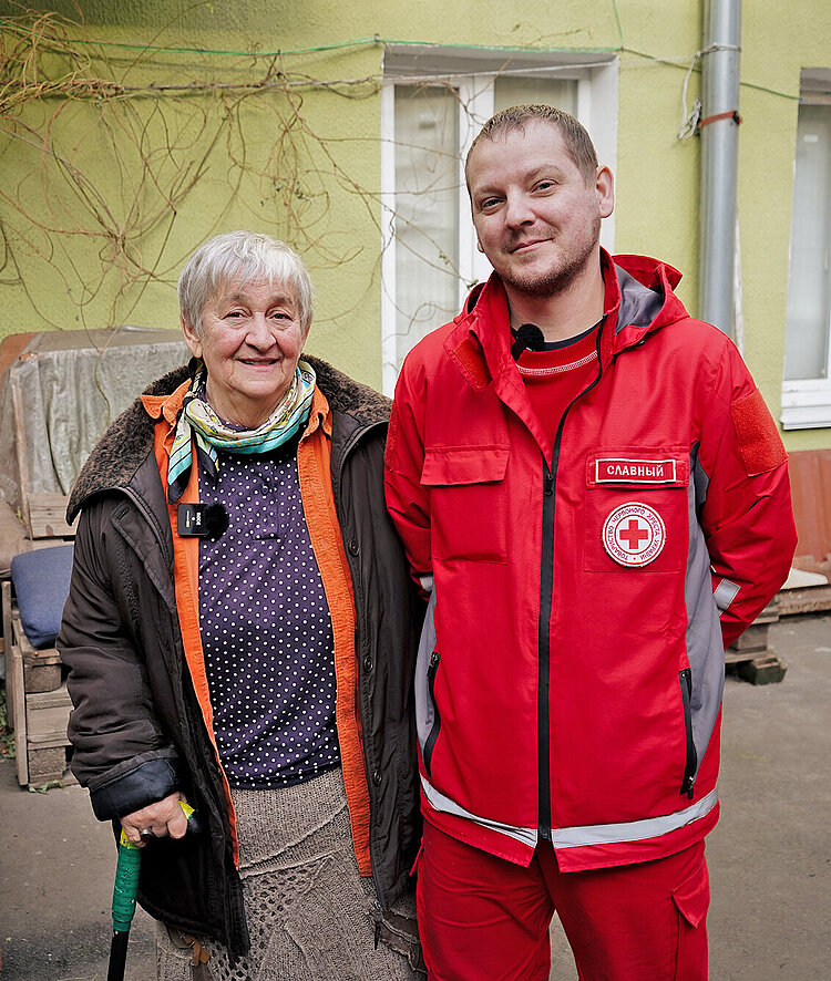 Auf dem Bild sieht man einen Helfer des Ukrainischen Roten Kreuzes, der rechts neben einer älteren Dame steht. Beide lächeln in die Kamera und stehen draußen.
