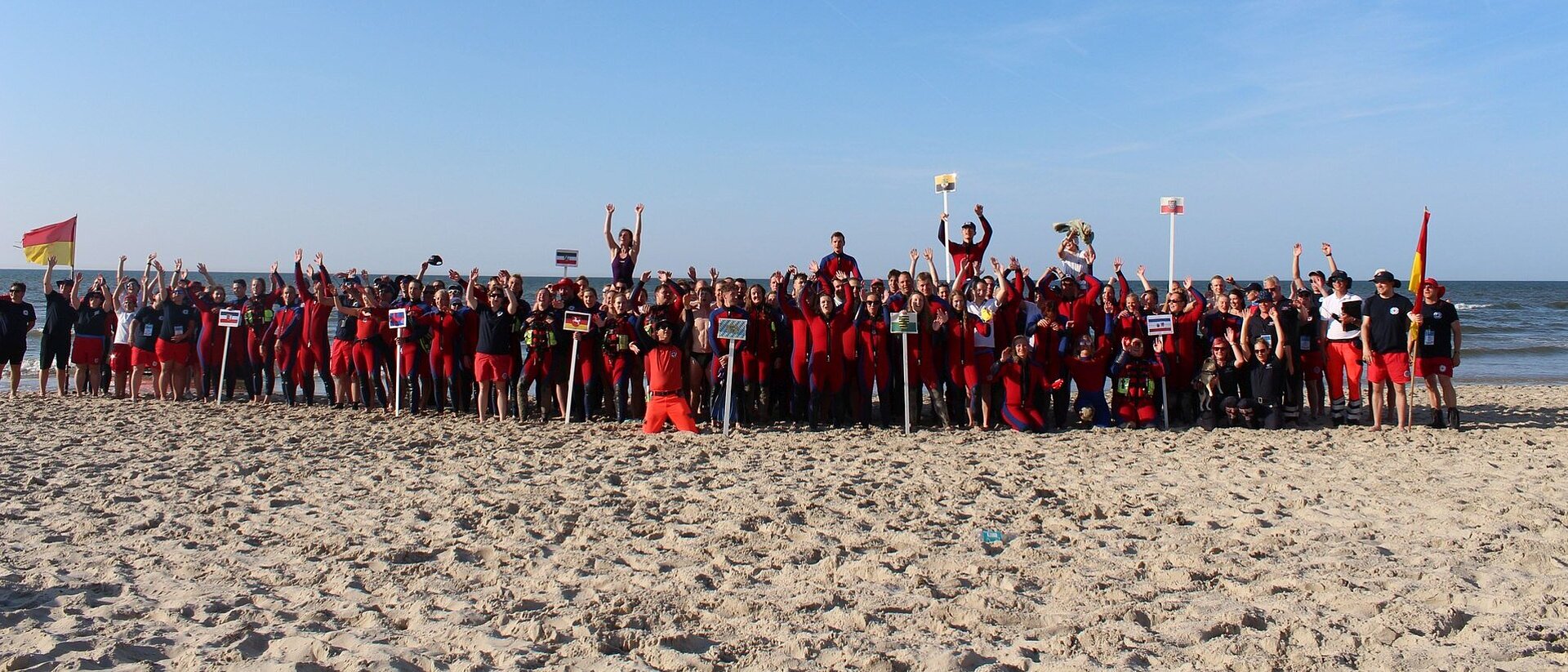 Am Strand von Wangerooge stehen alle Teilnehmenden und Beteiligten und halten die Arme hoch - Gruppenfoto am Strand mit über 100 Personen