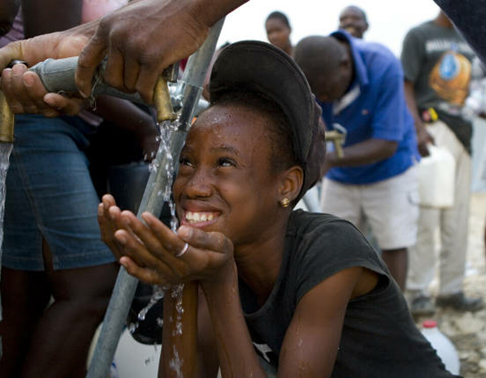 Foto: Mädchen lacht und trinkt Wasser aus einer provisorischen Wasserleitung.