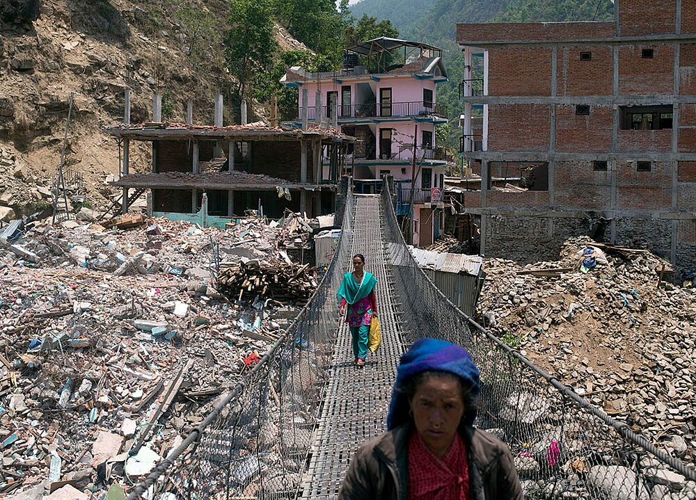 Menschen gehen über eine Hängebrücke, im Hintergrund die zerstörete Ortschaft Singati in Nepal