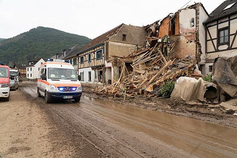 DRK-Einsatzfahrzeug unterwegs mit Blaulicht vor einem zerstörten Haus