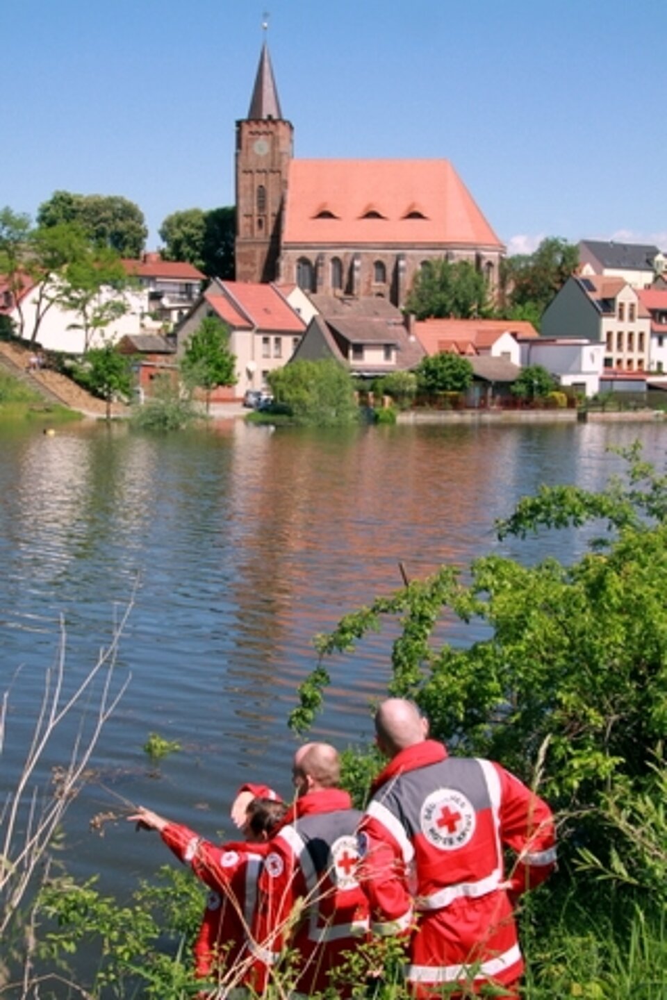 Foto: drei Männer in roten Rotkreuzjacken zeigen auf die Hochwasser führende Oder; auf der anderen Seite des Ufers ein Kirchturm und Gebäude.