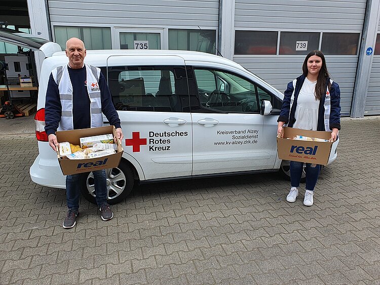 Ein DRK-Mitarbeiter und eine DRK-Mitarbeiterin stehen vor einem Lieferwagen und haben Kartons mit Lebensmitteln in der Hand