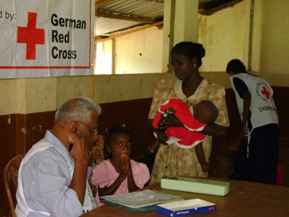 Foto: Ein Mädchen sitzt neben dem Mitarbeiter einer Gesundheitsstation an einem Tisch. Auf dem Tisch liegen Unterlagen. Neben dem Mädchen steht die Mutter. Sie hält ein Baby auf dem Arm.