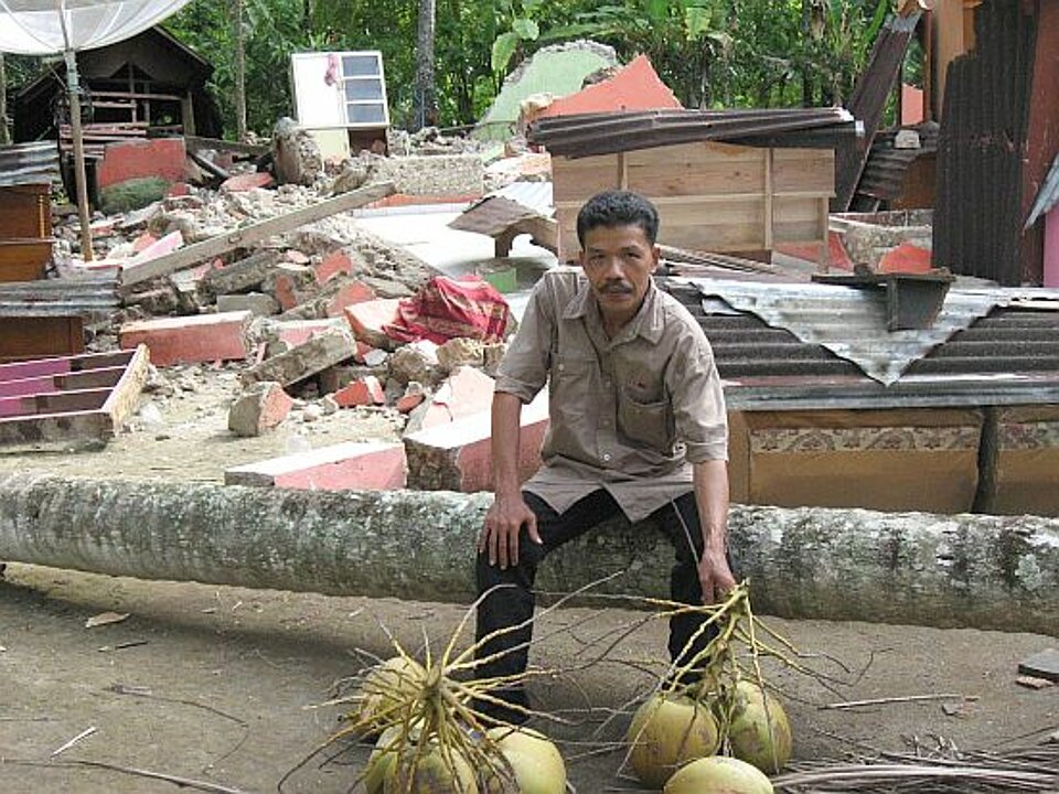 Foto: Ein asiatisch aussehender Mann sitzend vor einer Hausruine