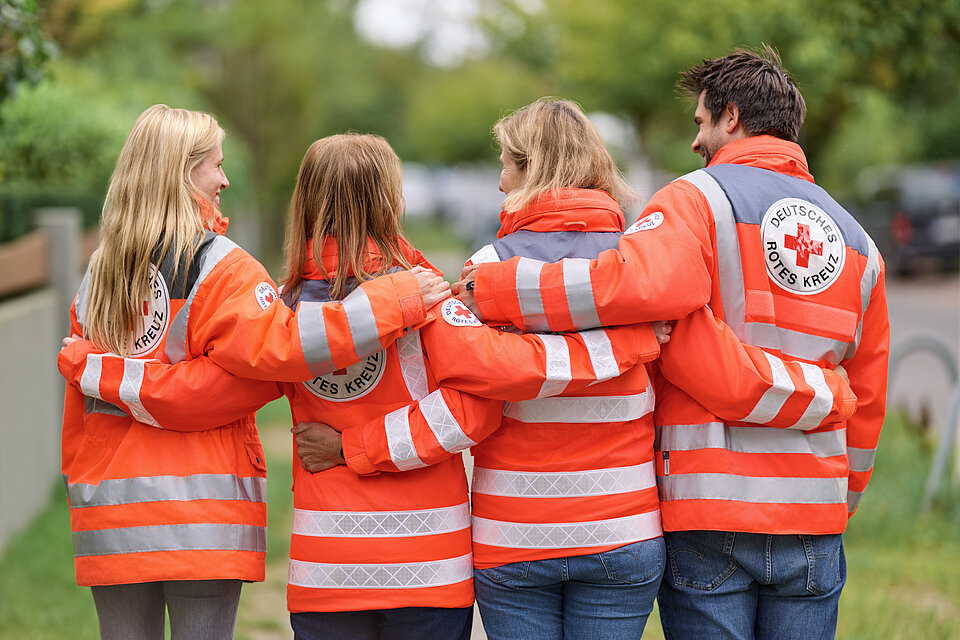 DRK-Helfende in Jacken in einer Reihe mit dem Rücken zur Kamera Arm in Arm