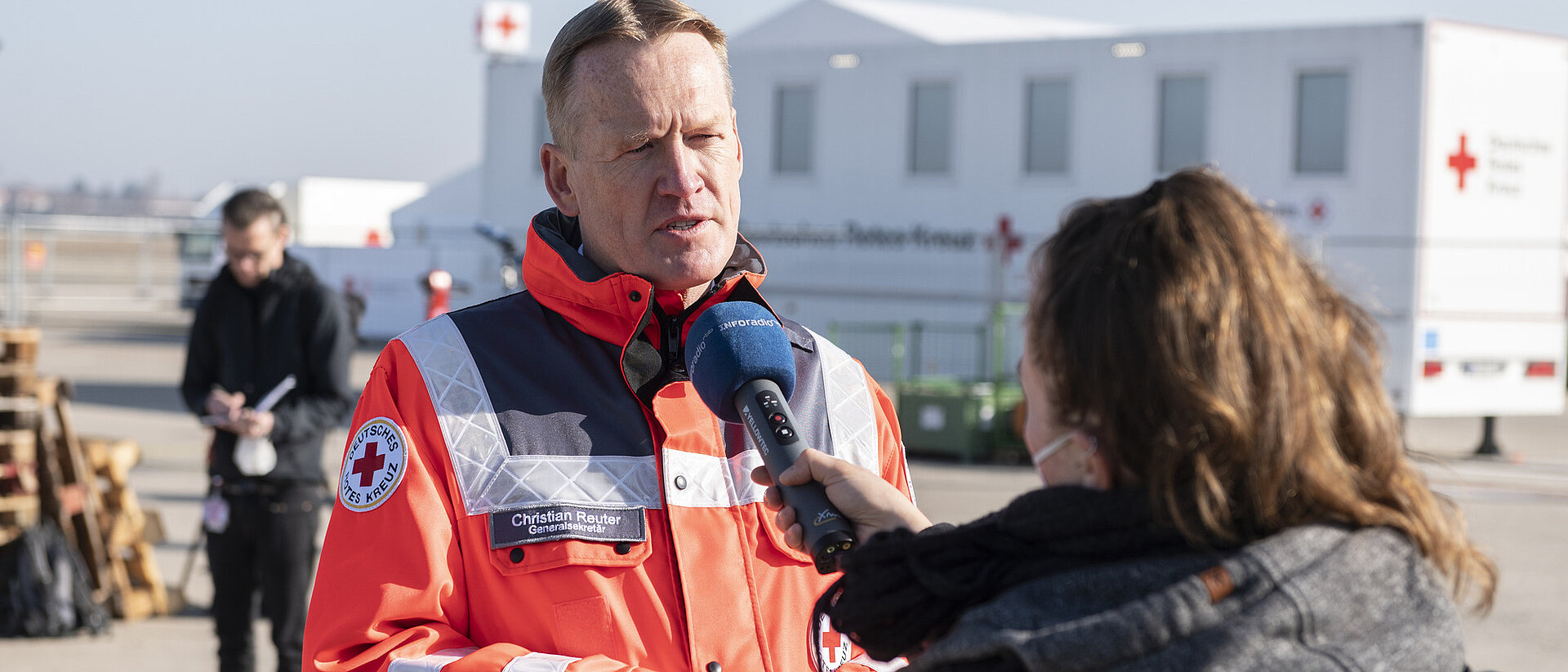DRK-Generalsekretär Christian Reuter steht in DRK-Einsatzkleidung im Freien und wird von einer Frau interviewt