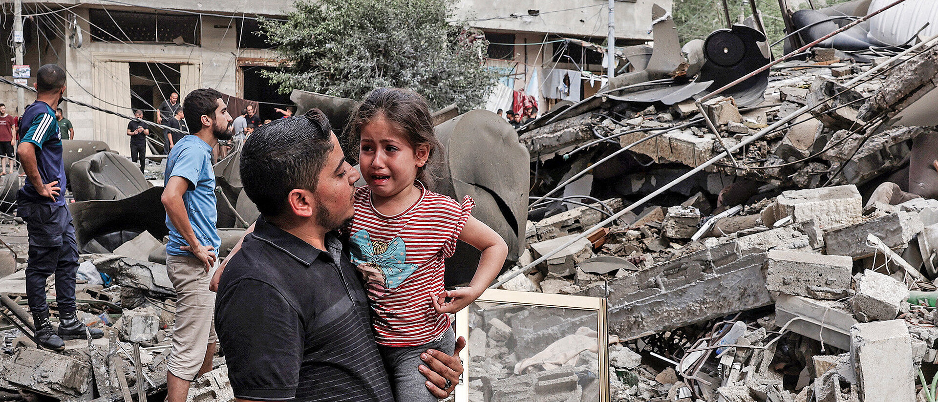 Ein Mann trägt ein Kind vor zerstörtem Gebaüde in Gaza am 7. Oktober 2023  