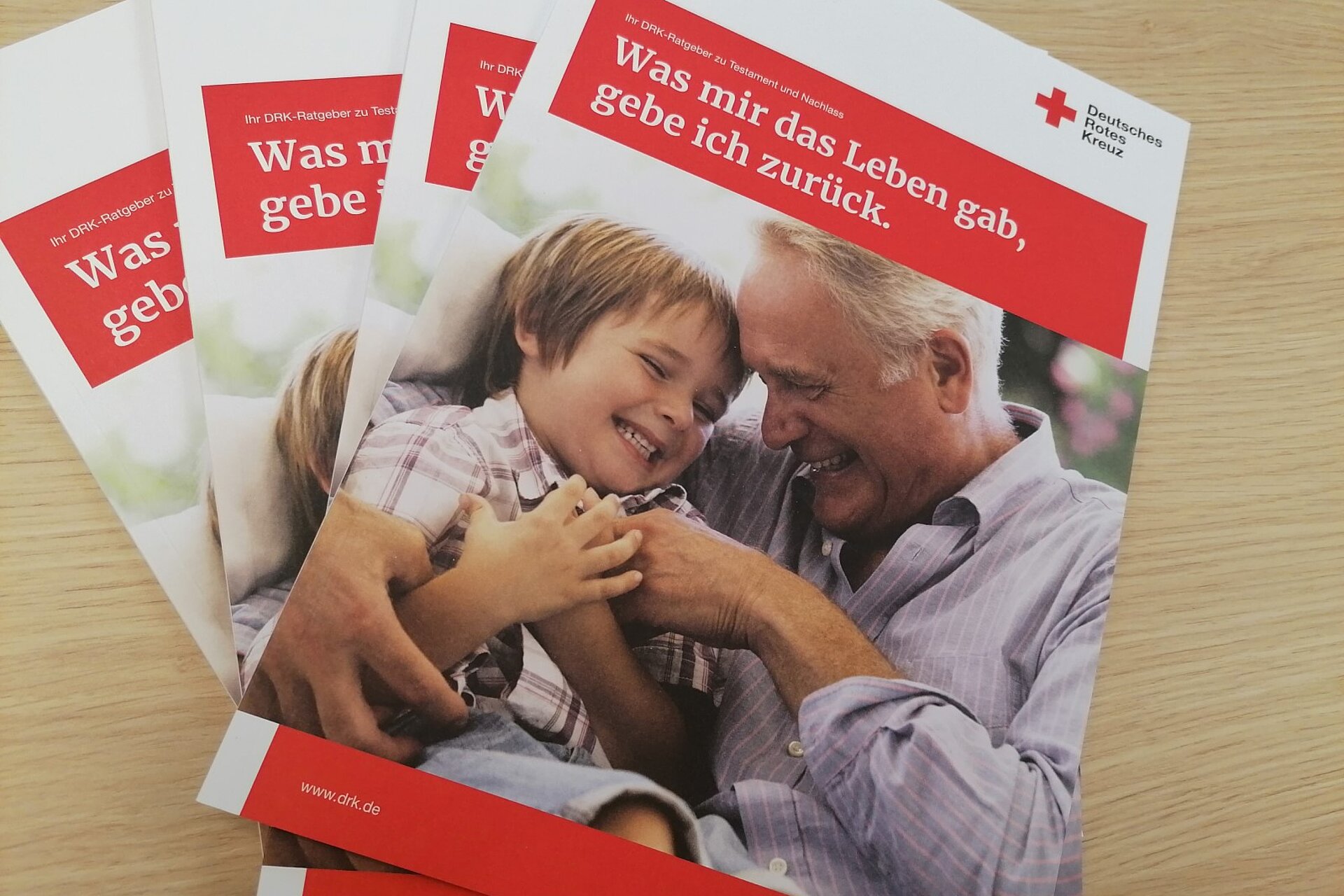 Vier Testamentratgeber vom Deutschen Roten Kreuz liegen übereinandergestapelt und leicht aufgefächert auf einem Tisch. 