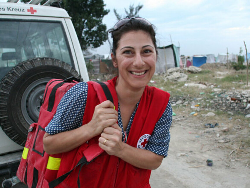 Foto: DRK-Mitarbeiterin vor einem DRK-Jeep und Trümmern in Haiti. 