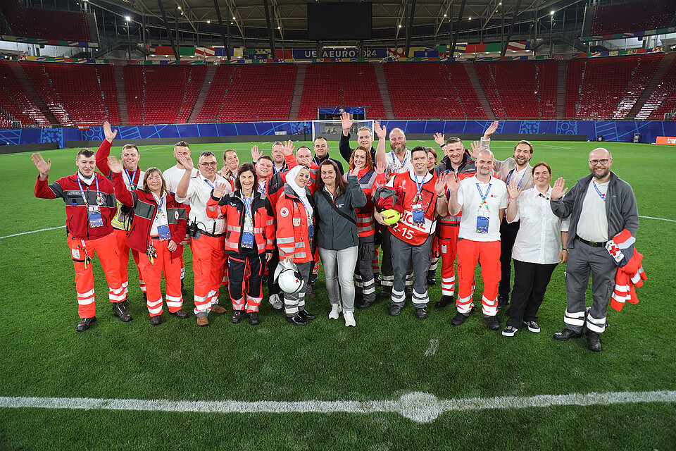 Eine große Gruppe von Rettungskräften des Deutschen Roten Kreuzes steht auf dem Spielfeld eines leeren Fußballstadions. Sie winken in die Kamera und tragen verschiedene DRK-Uniformen.