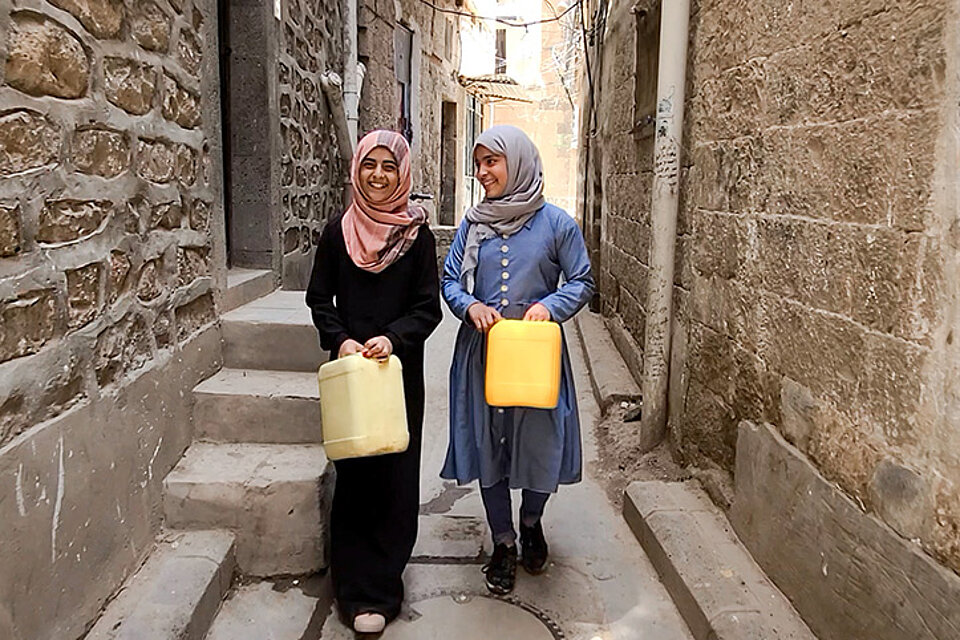 Zwei Mädchen mit Kopftüchern stehen in einer Gasse und halten Wasserkanister in der Hand