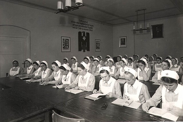 Schwesternhelferinnen an Bänken - Hakenkreuz an Wand