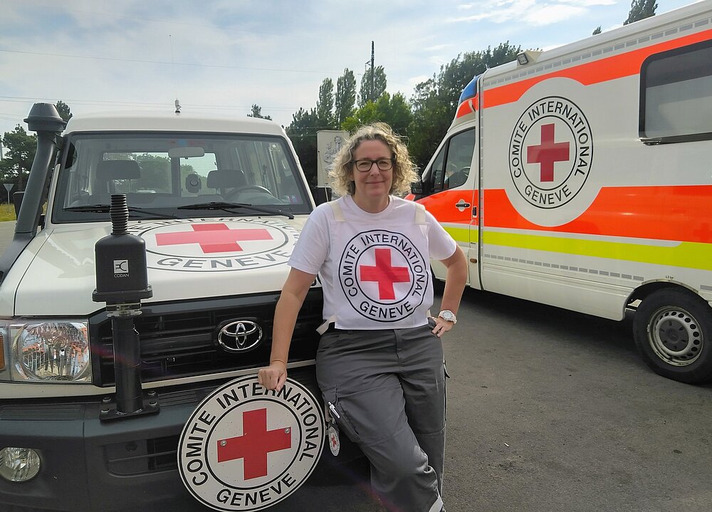 Eine Frau mit einem T-Shirt des Internationalen Komitees vom Roten KReuz steht vor einem IKRK-Fahrzeug. Im Hintergrund ein IKRK-Ambulanzfahrzeug.
