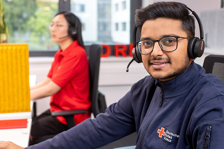 Ein junger Mann mit Headset und blauem Shirt mit DRK-Abzeichen lächelt den Betrachter an. Im Hintergrund sitzt eine Frau mit Headset.