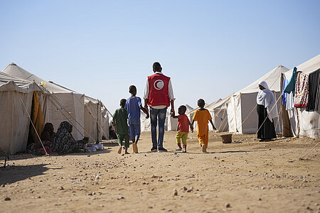 Helfender des Sudanesischen Roten Halbmonds geht mit Kindern an der Hand zwischen Zelten entlang
