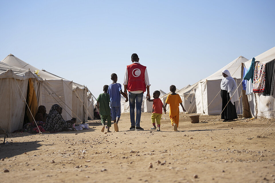 Helfender des Sudanesischen Roten Halbmonds geht mit Kindern an der Hand zwischen Zelten entlang