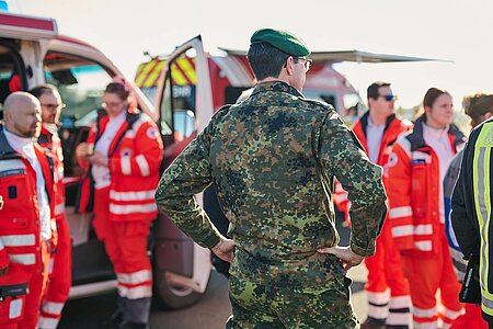 Im Vordergrund steht ein Mann in Bundeswehruniform mit dem Rücken zur Kamera im Freien. Im Hintergrund rechts und links sieht man mehrere Frauen und Männer in DRK-Dienstuniform.