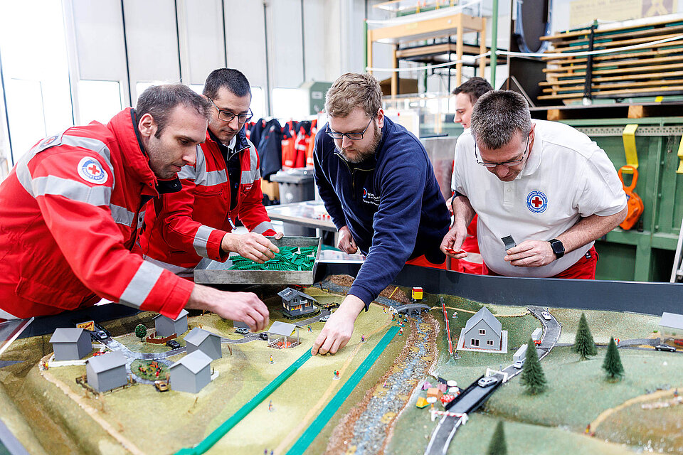 Vier Männer in DRK-Kleidung beugen sich über ein Tischmodell einer Siedlung 