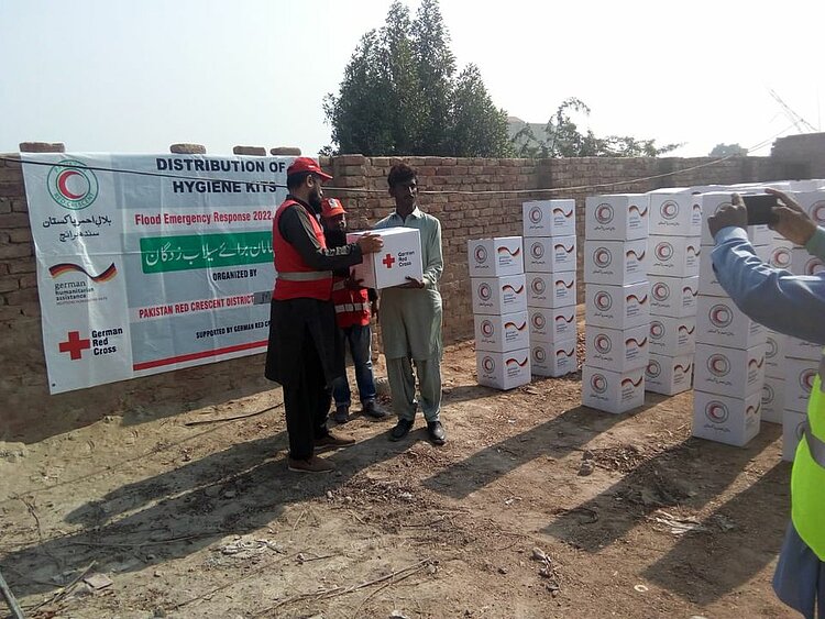 Mitarbeiter des Pakistanischen Roten Halbmonds überreichen einem Bewohner Matiayaris ein Hygieneset. Auf dem Karton ist das Logo des DRK zu sehen. 