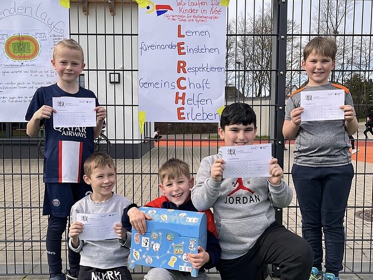 Auf dem Bild sind vier Schüler der Grundschule am Lerchenweg aus Monheim (NRW) zu sehen. Sie halten ihre ausgefüllten Spendenlauf-Zettel in den Händen und lächeln stolz in die Kamera. Sie tragen Sportsachen. Im Hintergrund hängt das Motto der Grundschule: Lernen, füreinander einstehen, respektieren, Gemeinschaft, helfen, erleben. 