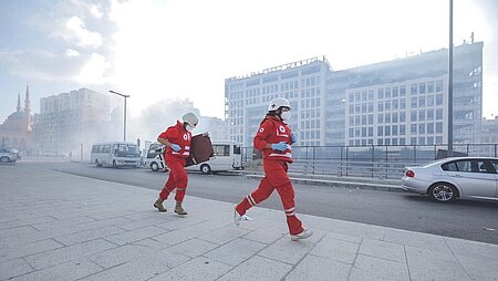Explosion in Beirut: Im Hintergrund sieht man Rauchschwaden und zwei Sanitäter des Libanesischen Roten Kreuzes laufen nach rechts 