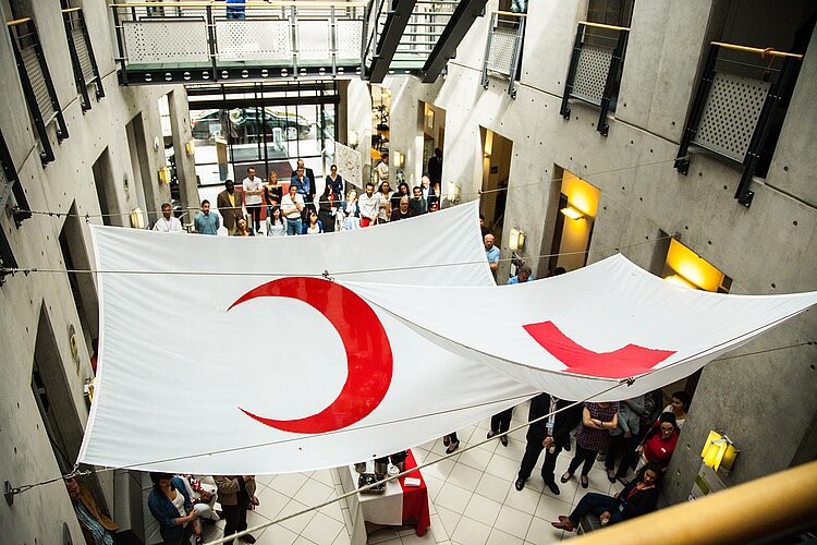 Blick von oben in das Gebäude der Internationalen Föderation der Rotkreuz- und Rothalbmondgesellschaften. Über den Köpfen der versammelten Menschen hängen zwei große Stoffbahnen mit den Symbolen des Roten Kreuzes und des Roten Halbmonds. 