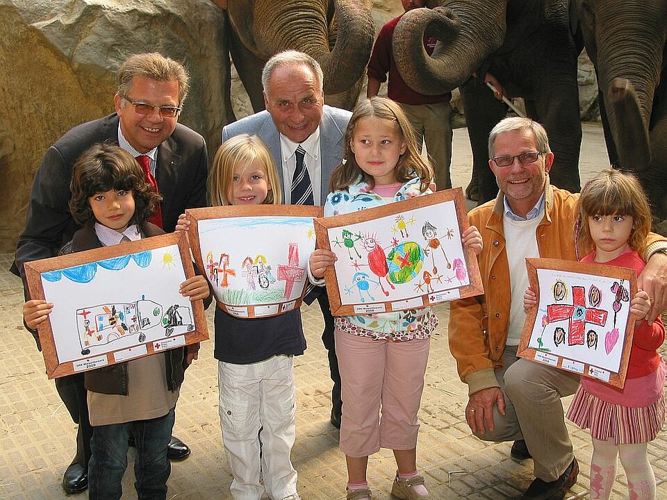 Foto: Vier Kinder halten selbst gemalte Bilder hoch, hinter ihnen drei ältere Herren
