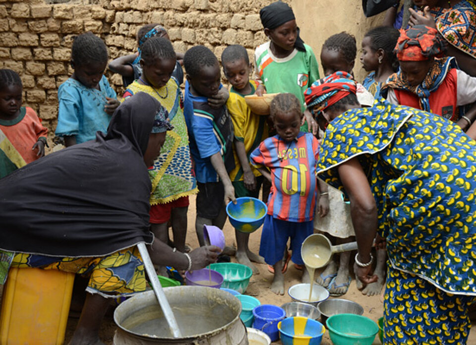 Afrikanische Kinder scharen sich um Schalen mit Haferbrei, die zwei Frauen füllen.