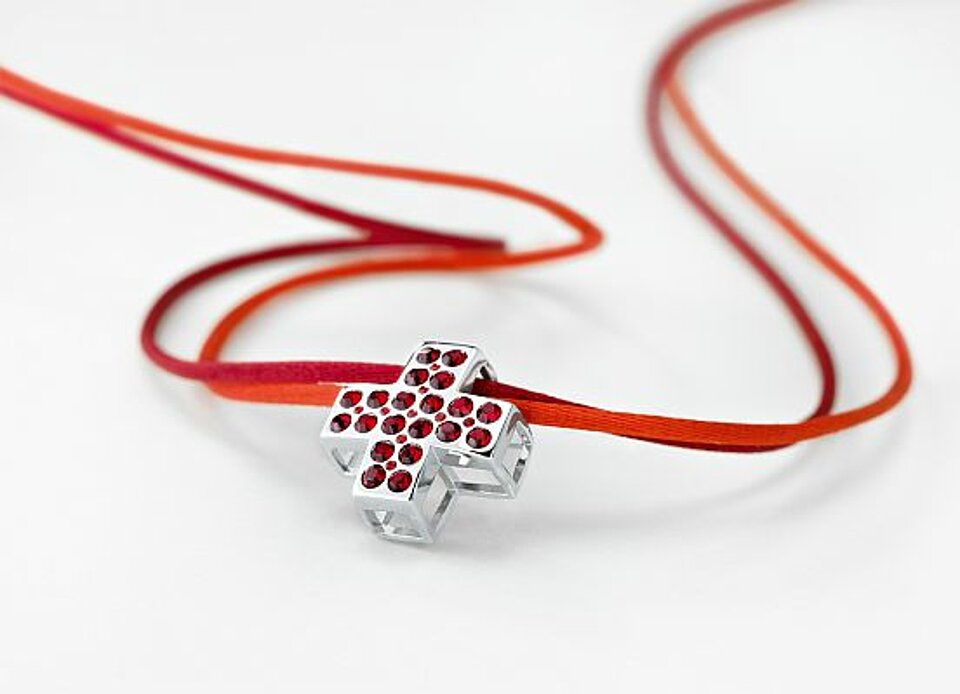 Foto: Das Rotkreuz-Collier von Pierre Lang: ein silbernes Kreuz, das mit kleinen roten Kristallen besetzt ist, aufgehängt an einem orangen und einem roten Textilband