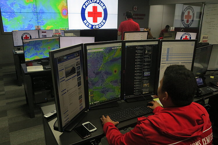 Ein Mitarbeiter des Philippinischen Roten Kreuzes arbeitet an mehreren Bildschirmen, auf denen Wetter- und Kommunikationsdaten angezeigt werden. Im Hintergrund sind weitere Mitarbeiter und ein großes Logo des Philippinischen Roten Kreuzes auf einem Bildschirm zu sehen.