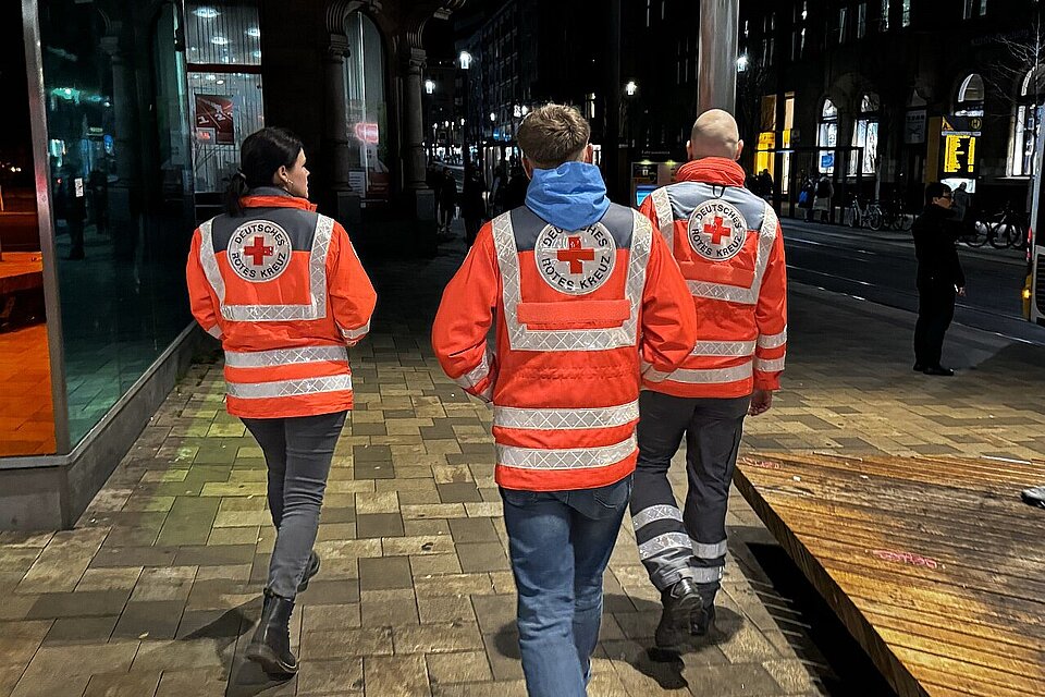 Drei DRK-Mitarbeiter mit Rotkreuzjacken gehen abends eine Straße entlang