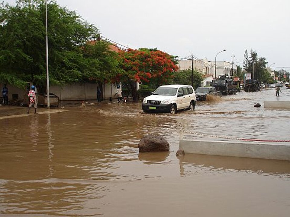 Foto: Ein überschwemmter Straßenzug mit umspülten Autos, im Hintergrund zwei Personen, die durch knietiefes braunes Wasser waten
