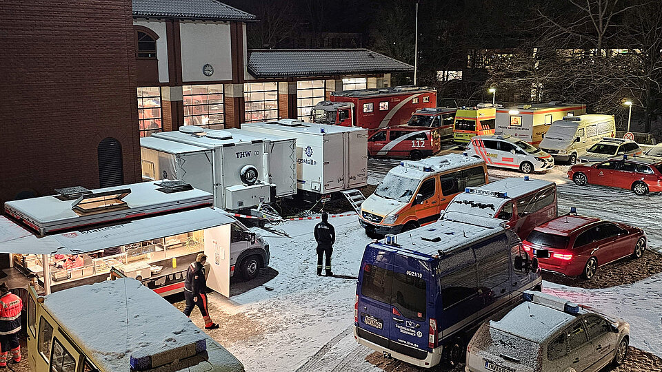 Parkplatz vor einer Feuerwache mit verschiedenen Einsatzfahrzeugen Nachtszene vor einer Feuerwache mit Schnee auf dem Boden und auf den Fahrzeugen. Mehrere Einsatzfahrzeuge von Feuerwehr, Rettungsdienst, Polizei und THW stehen dicht beieinander während Einsatzkräfte zwischen ihnen gehen und ein beleuchteter Verpflegungswagen auf einen laufenden Großeinsatz hinweist.