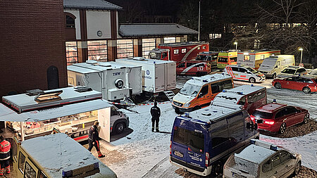 Nachtszene vor einer Feuerwache mit Schnee auf dem Boden und auf den Fahrzeugen. Mehrere Einsatzfahrzeuge von Feuerwehr, Rettungsdienst, Polizei und THW stehen dicht beieinander während Einsatzkräfte zwischen ihnen gehen und ein beleuchteter Verpflegungswagen auf einen laufenden Großeinsatz hinweist.