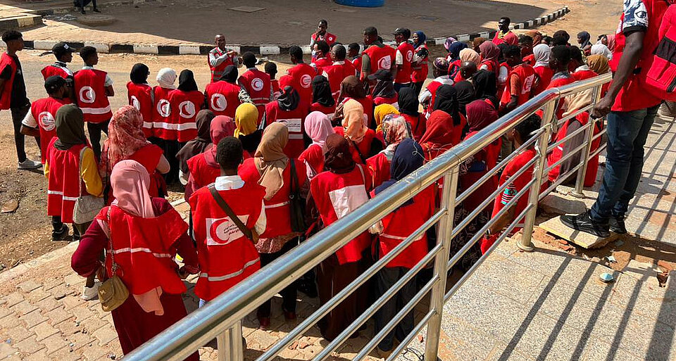 Eine große Gruppe von Freiwilligen und Mitarbeitenden des Sudanesischen Roten Halbmonds, erkennbar an ihren roten Westen mit dem Halbmond-Logo, versammelt sich im Freien. Die Mitarbeitenden, viele von ihnen mit Kopftüchern, stehen geordnet und hören einer Person zu, die vorne spricht.