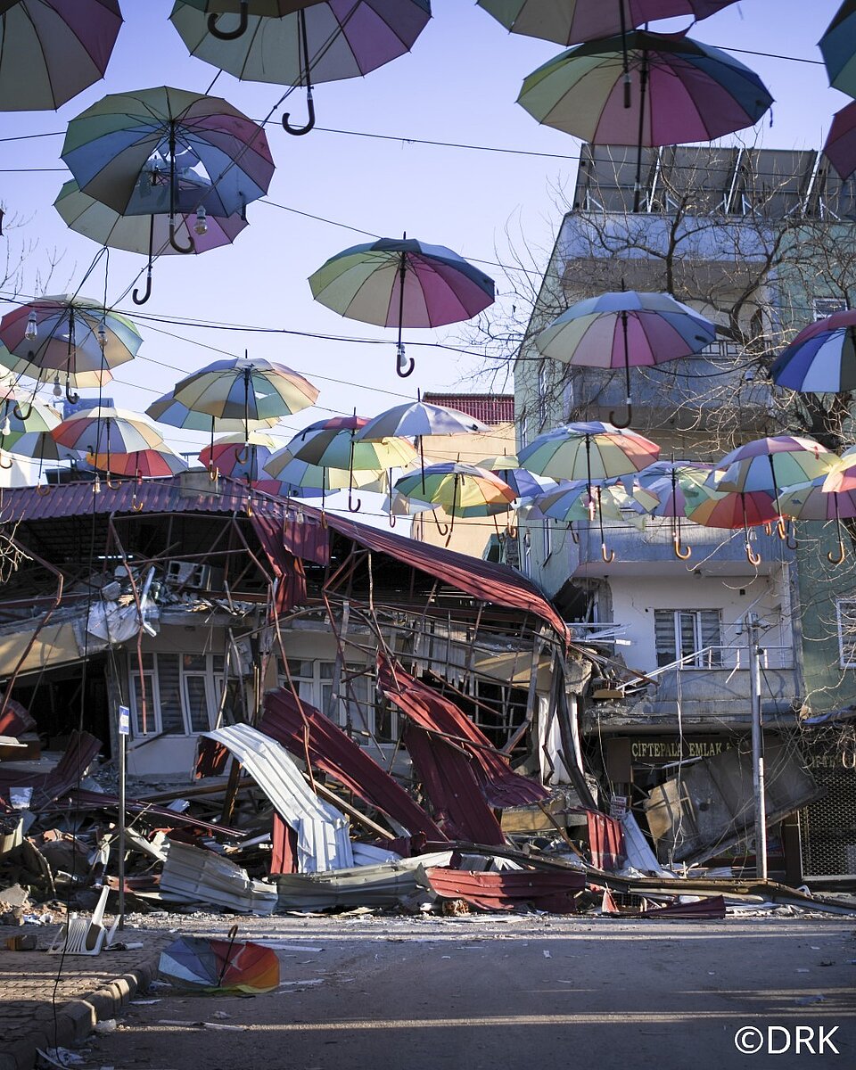 Das Bild zeigt zwei Häuser in einer Stadt der Türkei, die vom Erdbeben getroffen wurde. Das rechte, helle Haus hat vier Stockwerke und steht noch. Das linke Haus hingegen ist eingestürzt. Über der Straße, auf der die Häuser stehen, sind Regenschirme wie Girlanden gespannt. Sie sind geöffnet und haben Regenbogenfarben. Auf der Straße sind keine Menschen zu sehen, das Wetter ist sonnig.